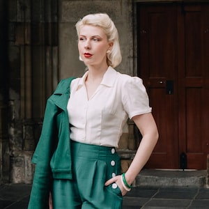 Pode incluir: Uma mulher em um traje de inspiração vintage. Ela usa uma blusa branca, calças verdes de cintura alta com botões brancos e uma jaqueta verde combinando sobre os ombros. Uma bolsa marrom e pulseiras verdes completam o look.