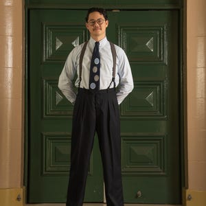 Puede incluir: Un hombre con camisa azul claro, tirantes negros y corbata azul marino con lunares blancos y azules. Lleva pantalones negros plisados y zapatos negros. Fondo de puerta verde.