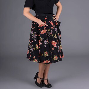 May include: A woman wearing a black blouse and a black and floral print skirt. She is standing in front of a gray background.