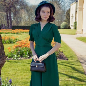 Può includere: Una donna con un vestito verde e un cappello bianco e nero tiene una borsa a mano nera. È in piedi in un giardino con un sentiero che conduce a un edificio sullo sfondo.