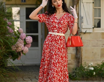 May include: A woman wearing a red floral dress with a white belt and a red handbag. She is wearing a red headband and white gloves.