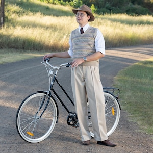 May include: A black bicycle with white tyres stands on a gravel road. The bicycle has a rear rack and reflectors. The person is wearing a brown hat, glasses, a patterned sweater vest, a white shirt, beige trousers, and brown shoes.