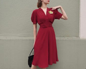 May include: A woman wearing a red vintage dress with a v-neckline and short sleeves. The dress has a fitted bodice and a flowing skirt. She is holding a black handbag and wearing a small flower brooch.