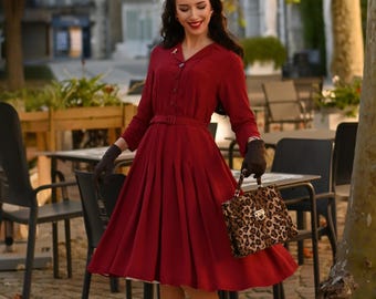 May include: A burgundy-colored dress with a button-down front, long sleeves, and a belted waist. The dress is paired with black gloves and a leopard-print handbag. A matching leopard-print headband is also visible.