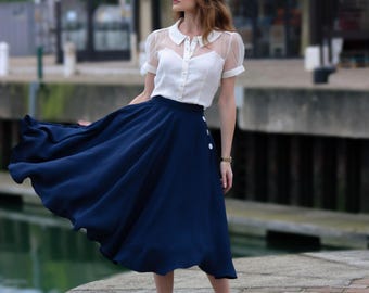 May include: A woman wearing a white blouse with sheer sleeves and a navy blue midi skirt with white buttons. The skirt is flowing in the wind.