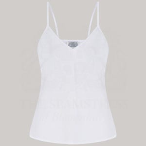 Puede incluir: Camisola blanca con finas tiras de espagueti y escote en V. La parte superior presenta un sutil patrón texturizado en la tela. Un pequeño emblema decorativo está centrado en el escote. Diseño simple y elegante.