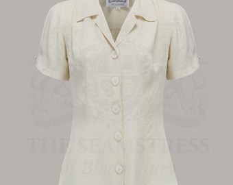 May include: A cream-colored short-sleeved button-down blouse with a pointed collar and a subtle tonal pattern. The garment features five round buttons down the center front and a label at the neckline that reads The Seamstress of Bloomsbury.