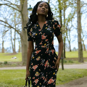 May include: A woman wearing a black floral dress with a V-neckline and short sleeves. The dress has a cinched waist and a flowing skirt.