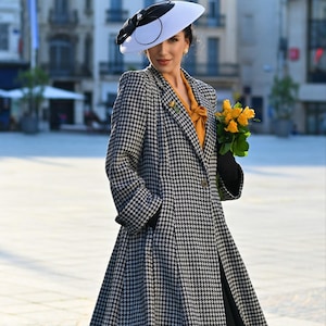 Könnte beinhalten: Eine Frau trägt einen schwarz-weißen Hahnentrittmantel, einen weißen Hut mit schwarzer Schleife und eine gelbe Bluse. Sie hält einen Strauß gelber Rosen. Der Mantel hat ein klassisches Design.
