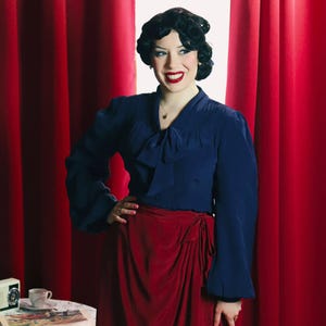 Puede incluir: Una mujer con una blusa azul marino con lazo y una falda roja de envoltura. Está de pie frente a una cortina roja.