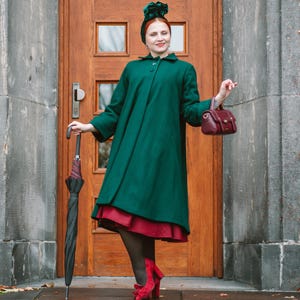 May include: A woman in a dark green coat, red dress, and heels, holding a burgundy handbag and a black umbrella. The background shows a wooden door and a stone wall. The outfit is vintage-inspired.