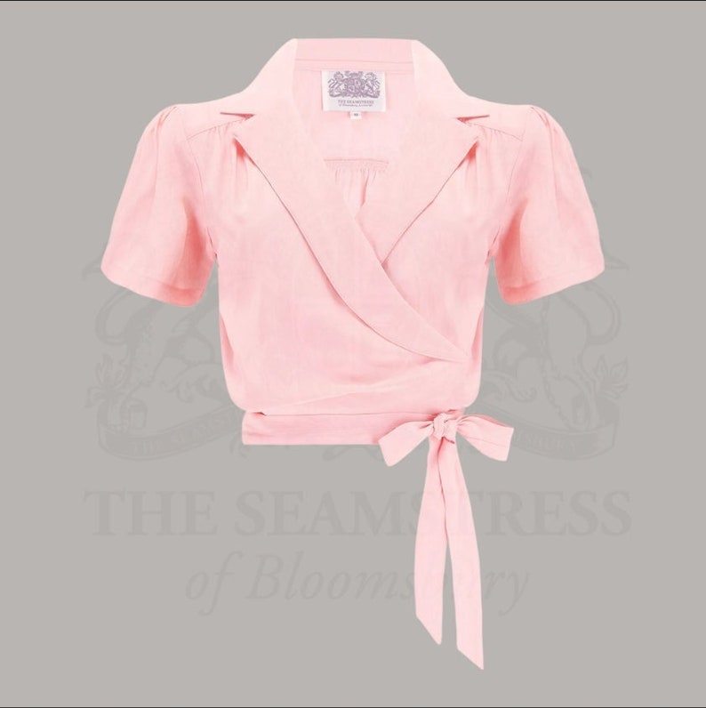 Op de afbeelding: Een lichtroze wikkel top met korte mouwen en een kraag. De top heeft een striksluiting in de taille en een klein label bij de halslijn. De tekst "THE SEAMSTRESS of Bloomsbury" is zichtbaar op de achtergrond.