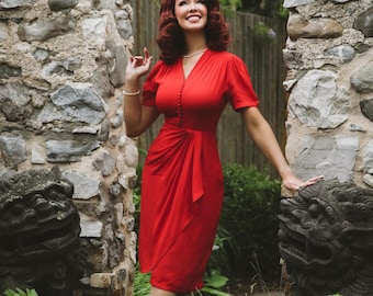 May include: A vibrant red knee-length dress featuring short sleeves, a V-neckline, and a vertical row of matching red buttons down the center bodice. The skirt includes a draped, wrap-style front detail that creates soft folds along the side.