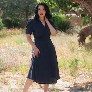 Può includere: Un abito midi blu navy scuro con maniche corte a sbuffo, scollo a V con corpetto a portafoglio e vita arricciata che scende in una gonna di media lunghezza. Il capo è realizzato in un tessuto a tinta unita con una leggera lucentezza.