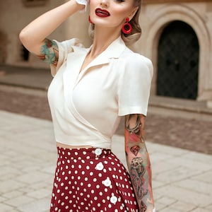 Op de afbeelding: Een vrouw in een vintage-ge&iuml;nspireerde outfit. Ze draagt een cr&egrave;mekleurige wikkel top en een bordeauxrode polkadot rok met witte knopen. Haar look wordt gecompleteerd met rode oorbellen, witte handschoenen en een retro kapsel.