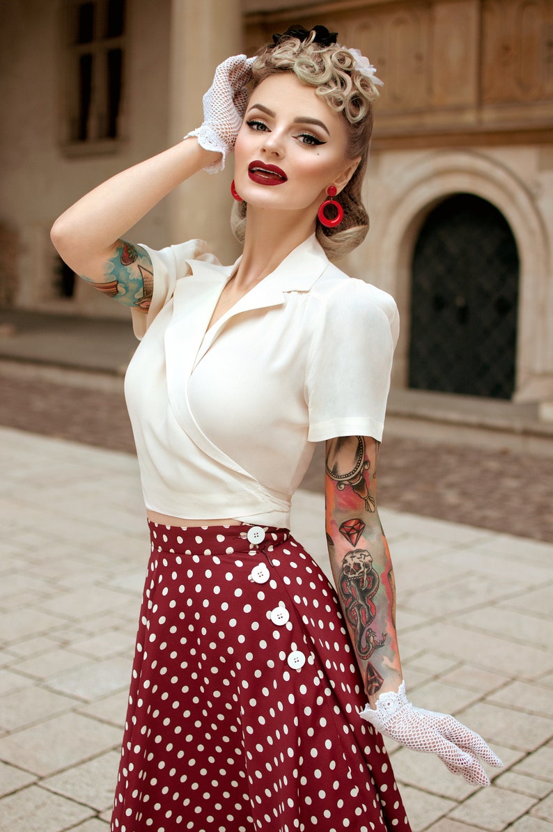 Op de afbeelding: Een vrouw in een vintage-ge&iuml;nspireerde outfit. Ze draagt een cr&egrave;mekleurige wikkel top en een bordeauxrode polkadot rok met witte knopen. Haar look wordt gecompleteerd met rode oorbellen, witte handschoenen en een retro kapsel.
