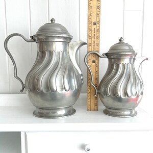 May include: Two antique pewter pitchers with lids. The larger pitcher is approximately 10 inches tall, and the smaller one is about 7 inches tall. Both have a fluted design and curved handles. They are displayed on a white surface.