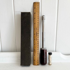 Puede incluir: Un conjunto de herramientas antiguas, que incluye una regla de madera, un destornillador con mango marr&oacute;n oscuro, un peque&ntilde;o vial de vidrio y una caja rectangular negra. La regla mide hasta 30 cm.