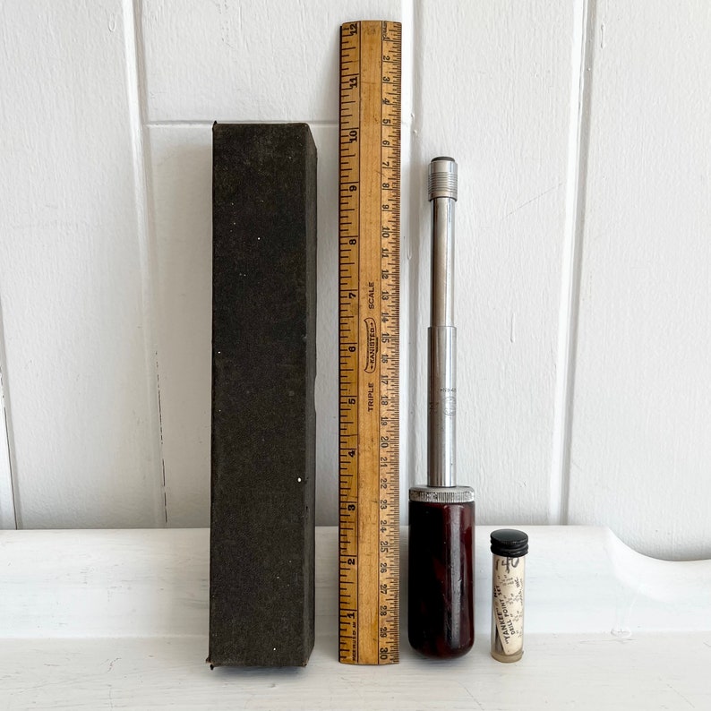 Puede incluir: Un conjunto de herramientas antiguas, que incluye una regla de madera, un destornillador con mango marr&oacute;n oscuro, un peque&ntilde;o vial de vidrio y una caja rectangular negra. La regla mide hasta 30 cm.