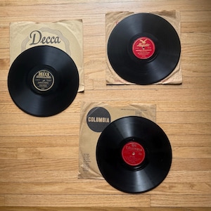 May include: Three vintage vinyl records on a wooden surface. Two records are in paper sleeves with the "Decca" and "Columbia" logos. The third record is in a paper sleeve with the "Majestic" logo. The records are black with colorful labels.