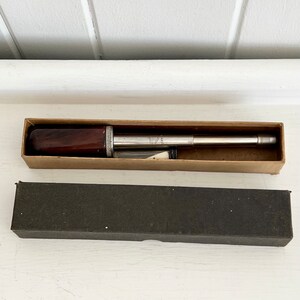 Puede incluir: Una herramienta vintage de plata y madera, posiblemente un instrumento de medici&oacute;n, se muestra en una caja de cart&oacute;n abierta. La herramienta tiene un mango de madera marr&oacute;n oscuro y un cuerpo de metal plateado. Debajo hay una segunda caja negra cerrada.