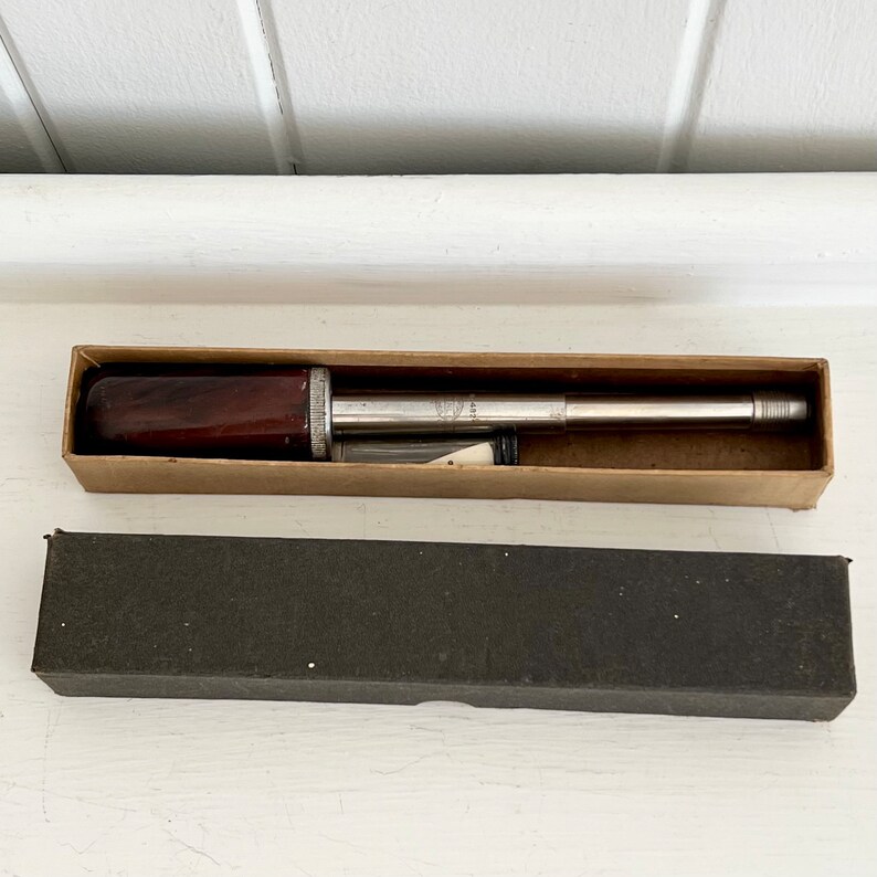 Puede incluir: Una herramienta vintage de plata y madera, posiblemente un instrumento de medici&oacute;n, se muestra en una caja de cart&oacute;n abierta. La herramienta tiene un mango de madera marr&oacute;n oscuro y un cuerpo de metal plateado. Debajo hay una segunda caja negra cerrada.