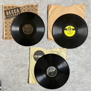 May include: Three vintage vinyl records are displayed, each with a black disc and a paper sleeve. One record is labeled "DECCA RECORDS", another "M-G-M", and the third "FM". The records are on a gray surface.