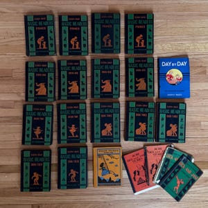 May include: A collection of vintage children's books, including "Basic Readers" in various volumes, "Health Stories," and "More Dick and Jane Stories." The books feature green, blue, and orange covers with illustrations of children and educational themes. The books are arranged on a wooden surface.