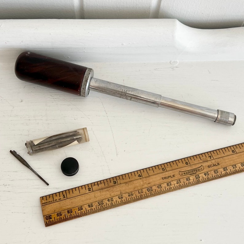 Puede incluir: Un destornillador vintage de metal y madera con un mango marr&oacute;n oscuro, un tubo de pl&aacute;stico transparente con peque&ntilde;as piezas de metal, una tapa negra y una regla de madera con medidas en pulgadas. La regla est&aacute; marcada con la palabra "TRIPLE".