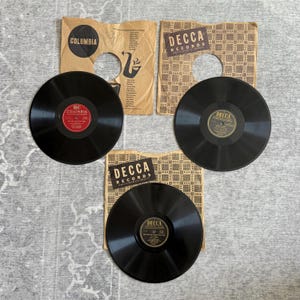 May include: Three vintage vinyl records with their sleeves. Two records are labeled "DECCA RECORDS" and one is labeled "COLUMBIA". The records are black with labels in red, gold, and black. The sleeves are tan with printed text and patterns.