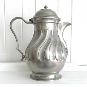 May include: A vintage silver-colored metal pitcher with a lid and handle. The pitcher has a fluted design on the body and a curved handle. The lid is topped with a small finial. The pitcher is likely used for serving beverages.