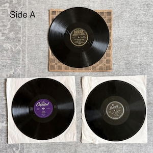 May include: Three black vinyl records are displayed. One record is in its original sleeve, labeled "DECCA." Two additional records are in white paper sleeves. One is labeled "Capitol" and the other "Mercury." The text "Side A" is visible.