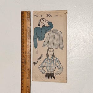 May include: A vintage sewing pattern envelope labeled 923, 20c, and size 17, featuring illustrations of long-sleeved button-down shirts. A wooden ruler is placed alongside the envelope, showing a length of approximately 12 inches.