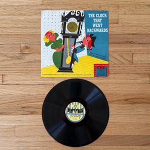 Op de afbeelding: Een vintage vinylplaat en albumhoes met de titel "The Clock That Went Backwards". De albumhoes heeft een kleurrijke illustratie van een klok en personages. De plaat is zwart met een geel en blauw label.