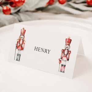Könnte beinhalten: Eine weiße Tischkarte mit einer Aquarellillustration eines Spielzeugsoldaten in roter Uniform. Der Name "HENRY" ist schwarz gedruckt. Die Karte liegt auf einem weißen Teller, mit roten Beeren und Grünzeug im Hintergrund.