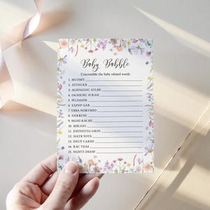 White baby shower game card featuring a pink, yellow, Lilac and peach wildflower border design on a white background The title Baby Babble is written in a script style font and below it a list of words baby related anagrams are written. The card is shown being held in a woman's hand