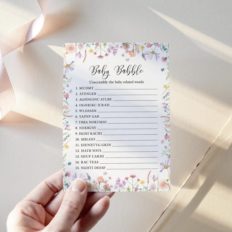 White baby shower game card featuring a pink, yellow, Lilac and peach wildflower border design on a white background The title Baby Babble is written in a script style font and below it a list of words baby related anagrams are written. The card is shown being held in a woman's hand