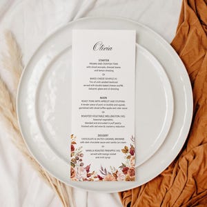 Peut inclure: Une carte de menu avec le nom "Olivia" en haut, ornée d'un motif floral dans les tons bordeaux, pêche et crème. Le menu est posé sur une assiette blanche, avec une seconde assiette en dessous. Un tissu marron et des plantes séchées sont en arrière-plan.