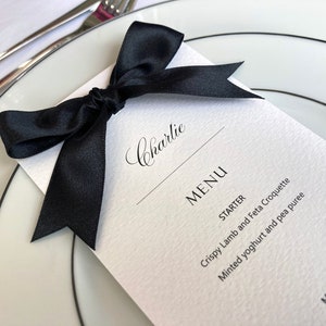 May include: A white place card with black text and a black satin ribbon tied in a bow. The text reads "Charlie" and "MENU". The menu includes "STARTER: Crispy Lamb and Feta Croquette, Minted yoghurt and pea puree" and "MAIN: fit duck leg".