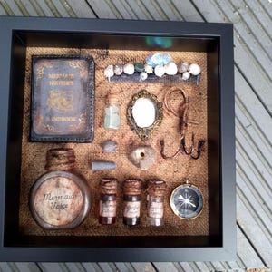 May include: A framed shadow box with a nautical theme. It includes a book titled "Mermaid Hunter's Handbook", a bottle labelled "Mermaid Voice", several small bottles with labels like "Mermaid Blood", "Algal Poison", and "Sea Serpent Tears", a compass, a mirror, a comb, seashells, and other nautical items.