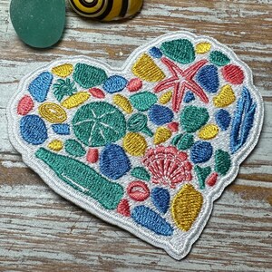Könnte beinhalten: Gestickter herzförmiger Aufnäher mit Strandmotiv. Das Design umfasst einen roten Seestern, Muscheln und Meeresmotive in Blau, Gelb und Grün, alle mit weißen Nähten umrandet.
