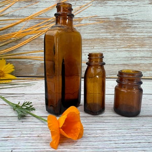 May include: Three amber glass bottles of varying sizes on a whitewashed wood surface. The bottles are empty and have a vintage look. A single orange poppy flower lies in the foreground.