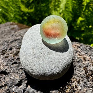 Op de afbeelding: Een bolvormige, doorschijnende knikker van zeeglas met een matte textuur en interne gele en oranje wervelingen, rustend op een gladde, lichtgrijze, afgeronde steen. De knikker vertoont een zachte, diffuse gloed door het zonlicht.