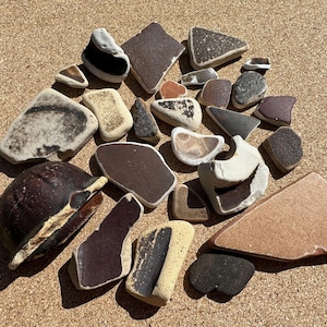 May include: A collection of sea glass pieces in various shapes and colors, including brown, white, and tan. Some pieces have rounded edges, while others are more angular. The pieces are arranged on a textured, light brown surface.