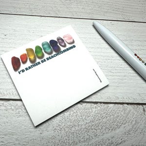 May include: A white notepad with the text "I'D RATHER BE BEACHCOMBING" and a row of colorful, gem-like shapes. A white pen with a rainbow design is next to the notepad. The notepad and pen are on a light wood surface.