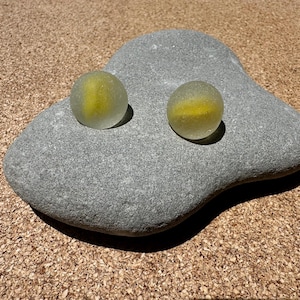 Op de afbeelding: Twee bolvormige glazen objecten met een matte, doorschijnende witte afwerking en een heldergele kern. Deze ronde voorwerpen zijn geplaatst op een gladde, grijze, onregelmatig gevormde steen, wat een duidelijk contrast tussen glas en steen toont.