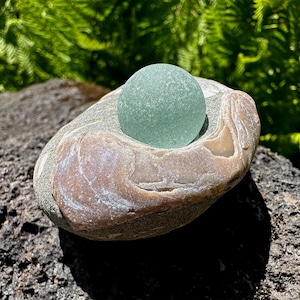 May include: A smooth, translucent, pale sea-foam green glass sphere rests securely within a natural indentation of a multi-colored, polished river stone. The stone features swirling patterns of tan, brown, and grey with a smooth, weathered texture.