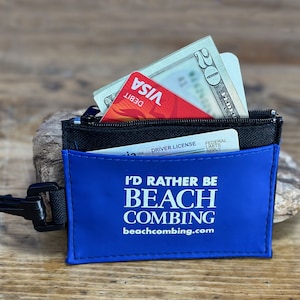 May include: A blue zippered pouch with a black strap and a carabiner clip. The pouch has the text "I'd Rather Be Beach Combing" and the website "beachcombing.com".