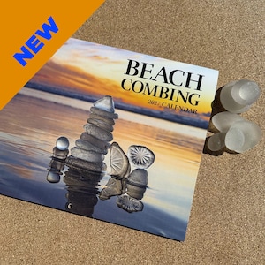 May include: A 2027 wall calendar titled BEACH COMBING featuring a photograph of stacked stones and sea glass on a beach at sunset. The calendar cover displays a tall cairn of stones, translucent glass pieces, and the text NEW in blue.