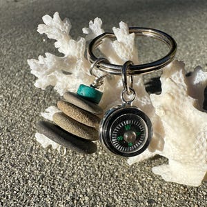 May include: A silver keychain with two charms. One charm is a small compass, and the other is a stack of small, smooth stones. The stones are in shades of gray and brown, with a turquoise bead at the top. The keychain is resting on a piece of white coral.